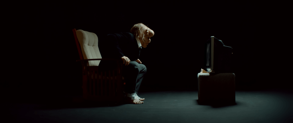 Ein barfüßiger Mann in Anzug lehnt sich in einem dunklen Raum gespannt aus einem Sessel vor einen kleinen Fernseher.