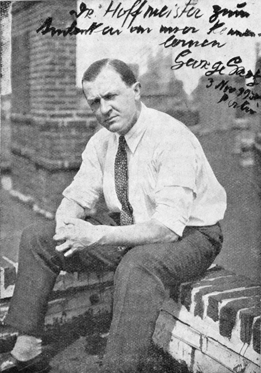 Schwarz-weißes Ganzkörperfoto von George Grosz; er sitzt auf einer Mauer vor Stadtkulisse, mit handschriftlicher Widmung.
