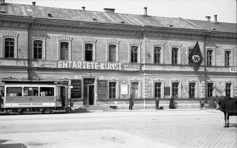 Schwarz-Weiß-Foto: Gebäude in Salzburg mit Banner „ENTARTETE KUNST“, Hakenkreuzfahne, Straßenbahn und Passanten davor.